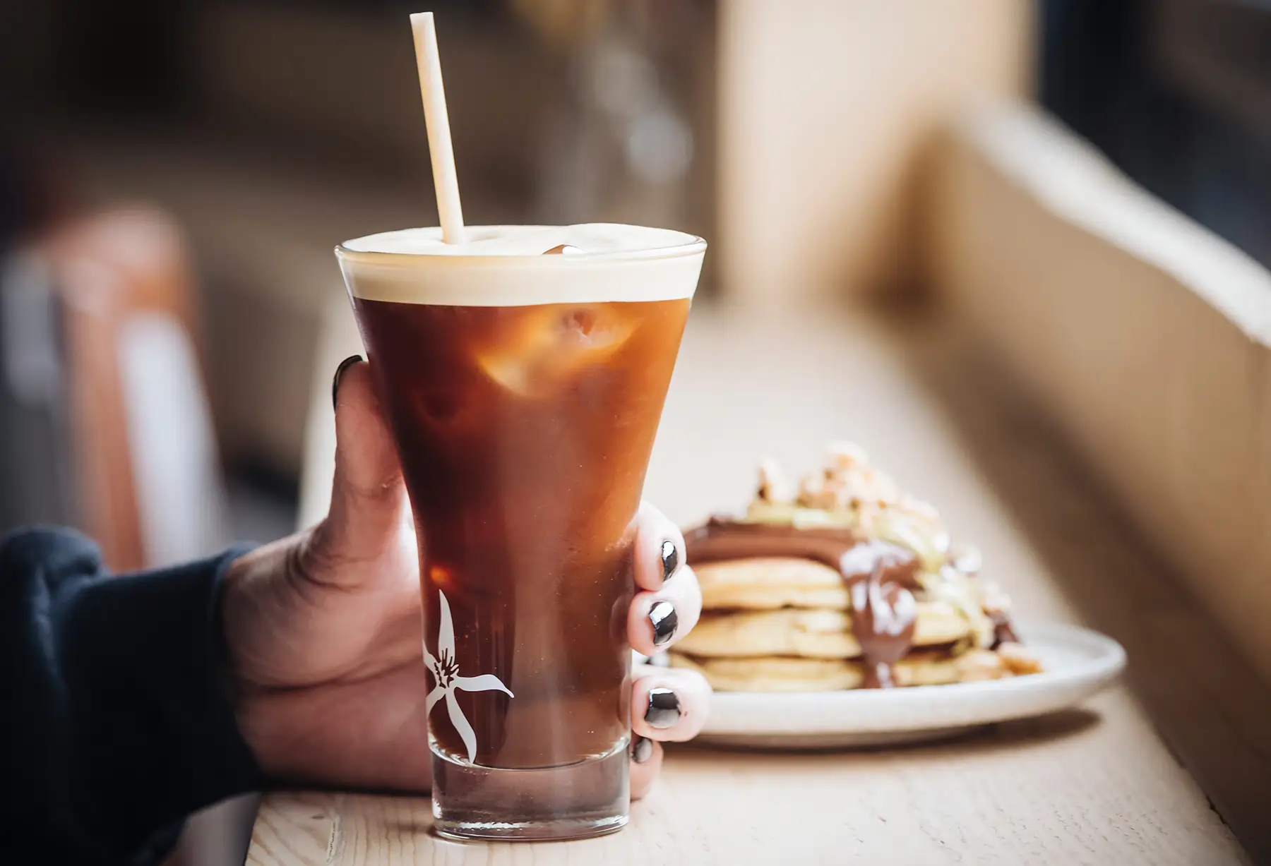 Hand holding cold brew coffee next to chocolate pancakes at Bread and Spread